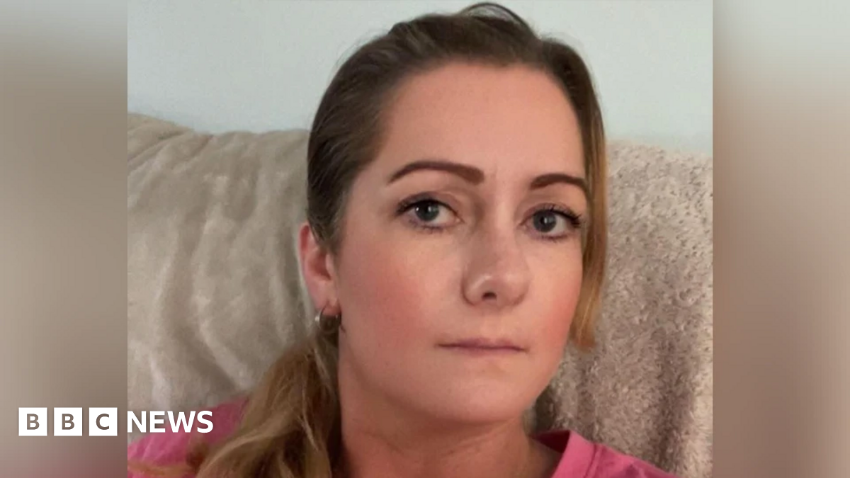 A white woman with dark hair and dark eyes lookiing at the camera, she is wearing a pink top