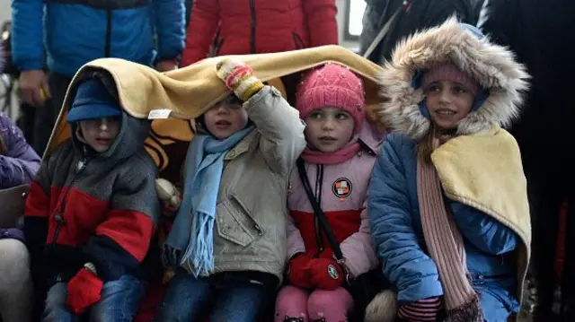 Four children wrapped up in winter coats sit under a yellow blanket