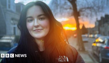 Kate Finlay, young woman smiling at camera in street, with sun shining in the background.