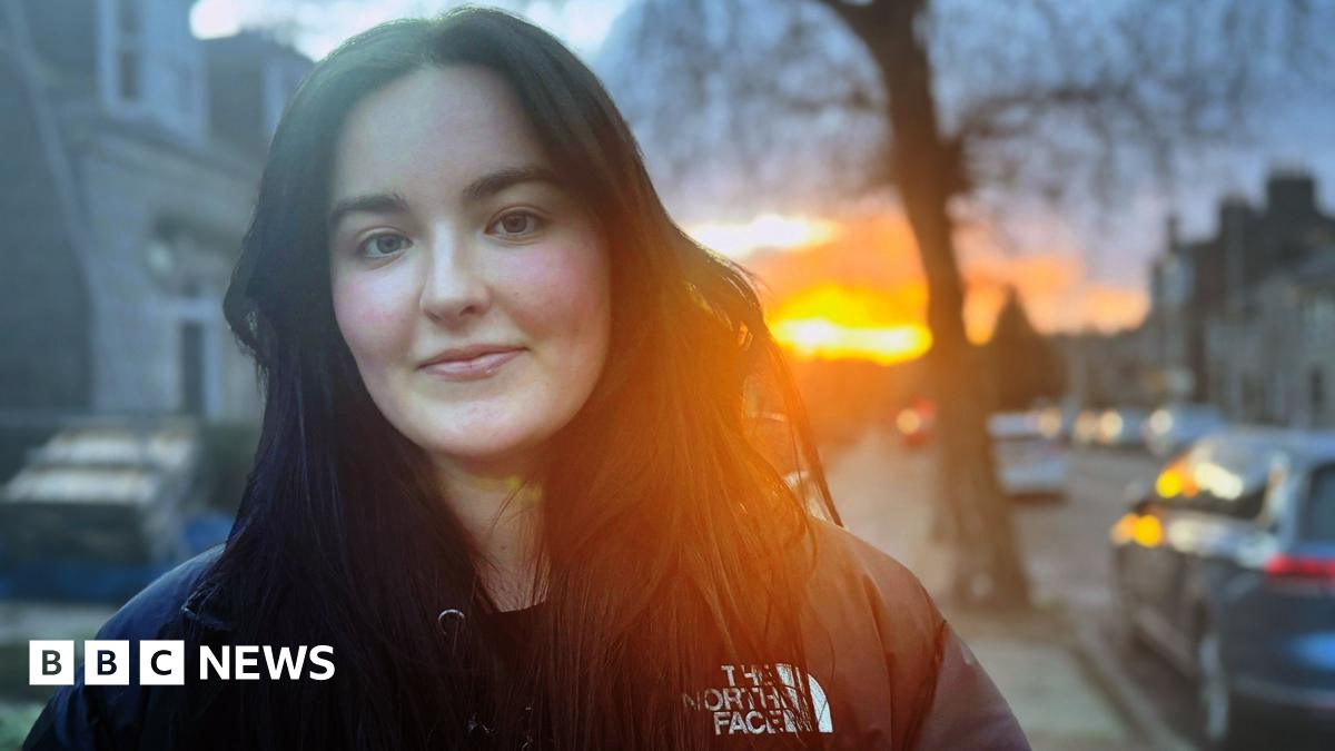 Kate Finlay, young woman smiling at camera in street, with sun shining in the background.