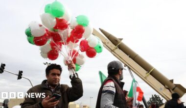 An Iranian man holds up a bunch of green, red and white balloons near a ballistic missile during celebrations marking the 47th anniversary of the Islamic revolution in Tehran, Iran (11 February 2026)
