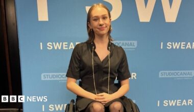 A woman in a wheelchair wearing a black dress in front of a promotional board for the film I Swear