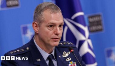 A clean-shaven man in a blue military uniform sits at a table in front of a Nato flag for a press briefing.