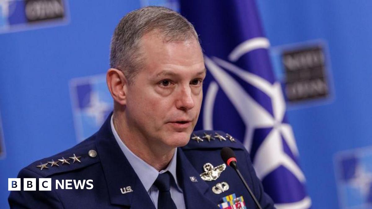 A clean-shaven man in a blue military uniform sits at a table in front of a Nato flag for a press briefing.