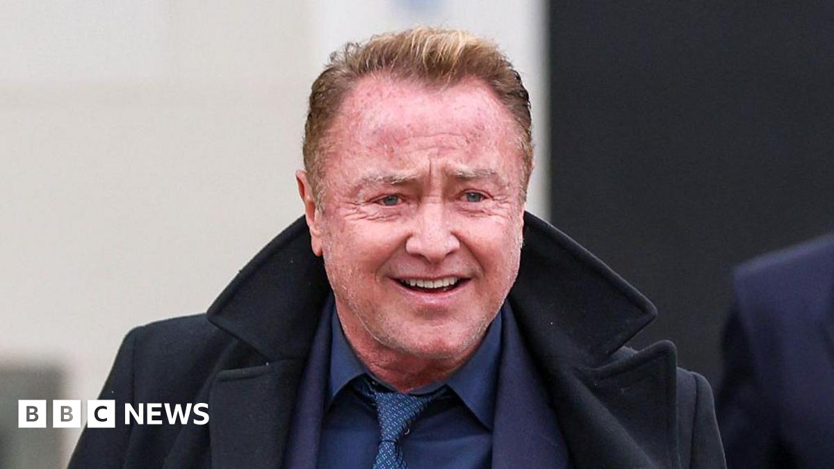 Michael Flatley. He has short reddish hair, wearing a navy suit and black coat.