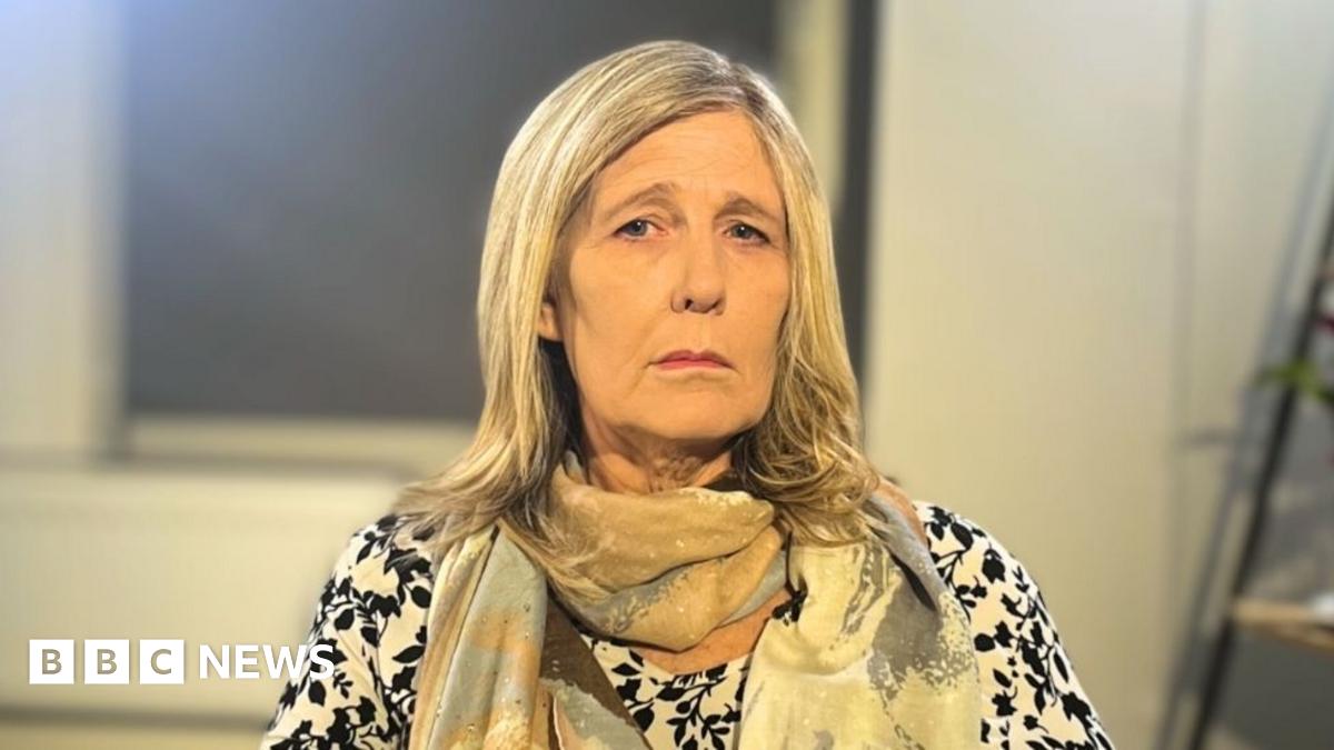 Vanessa is looking at the camera. She is wearing a black and white patterned long sleeve top and a brown, blue and cream scarf. She has shoulder length blonde hair. Behind her, there are shelves with plants on them and a window with blinds shut.