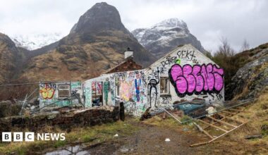 The cottage is a ruin and its white walls are covered in graffiti. Behind the ruins loom large high mountains.