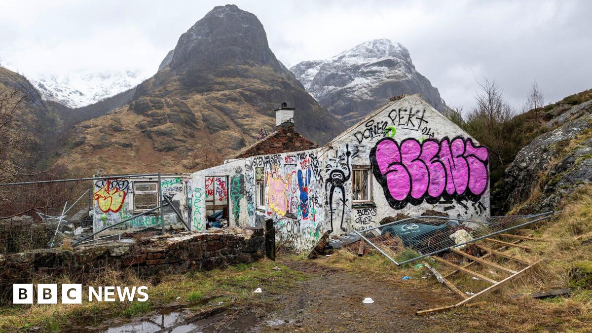 The cottage is a ruin and its white walls are covered in graffiti. Behind the ruins loom large high mountains.