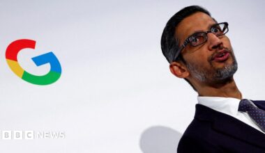 Google's rainbow colored 'G' logo on a white wall behind CEO Sundar Pichai, wearing a black suit, white shirt and purple neck tie.