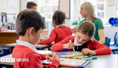 A close up view of a small group of school friends sitting in their classroom. Two pupils at the front of the image are drawing pictures on their whiteboards and a teaching assistant is supervising other students in the background.
