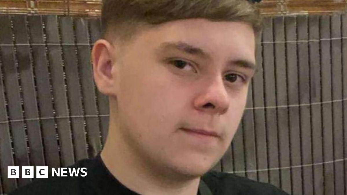 A teenage boy with dark hair. He is staring into the camera. A fence is behind him.