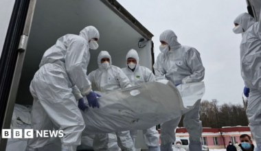 Officials in white overalls that cover their heads and wearing face mask carry a white stretcher presumably containing a body being unloaded from what looks like a truck
