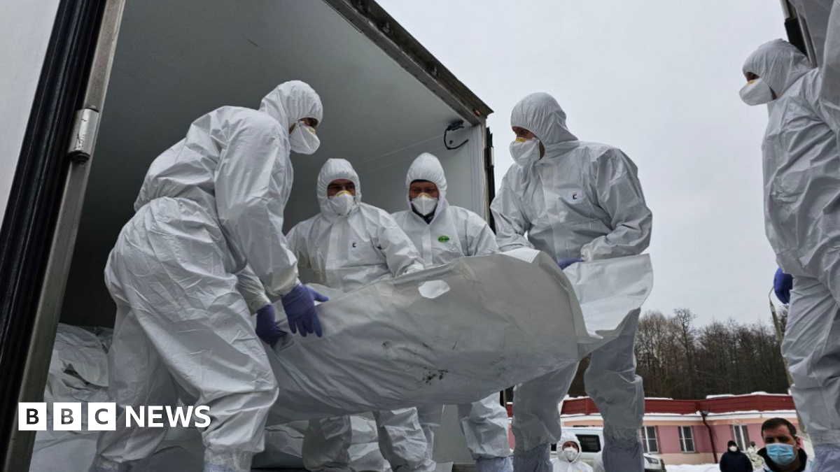 Officials in white overalls that cover their heads and wearing face mask carry a white stretcher presumably containing a body being unloaded from what looks like a truck