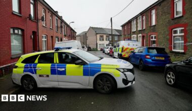 Street with terraced houses either side and cars parked alongside the road. A police car is parked across the road to prevent access and police tape has been placed across the road.