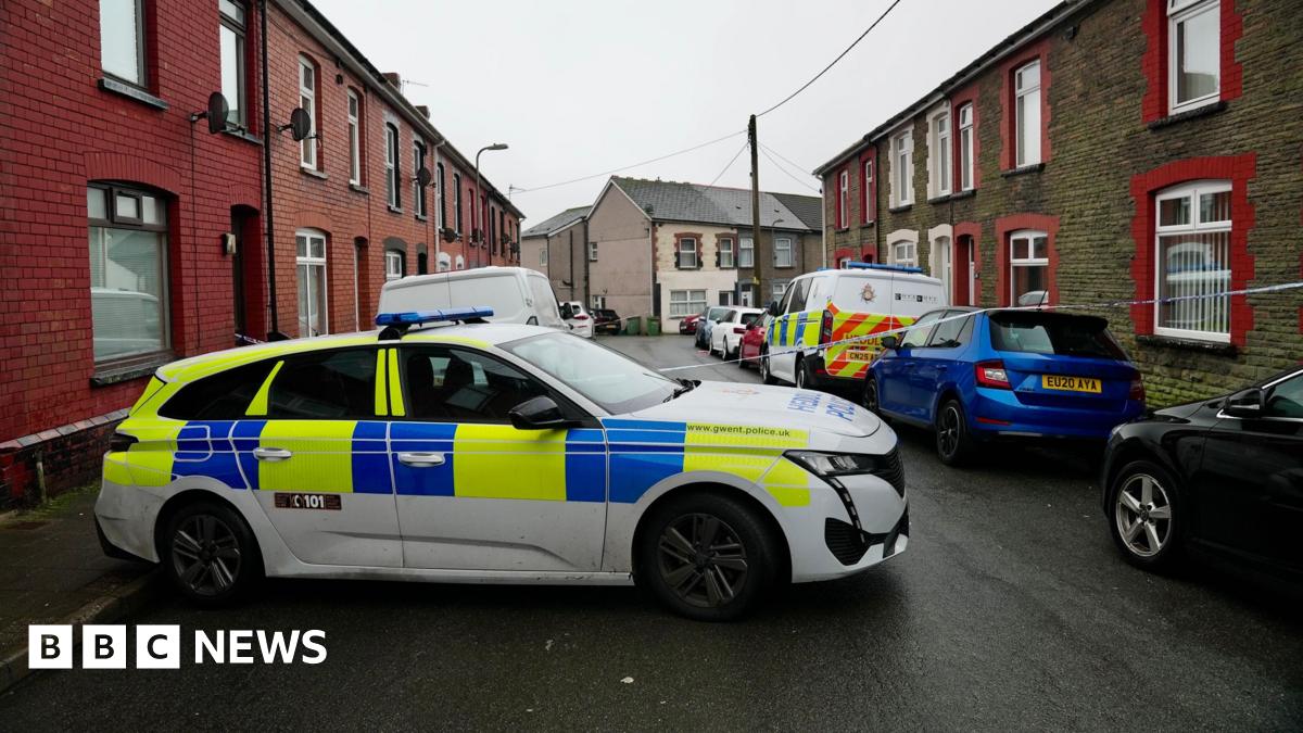 Street with terraced houses either side and cars parked alongside the road. A police car is parked across the road to prevent access and police tape has been placed across the road.