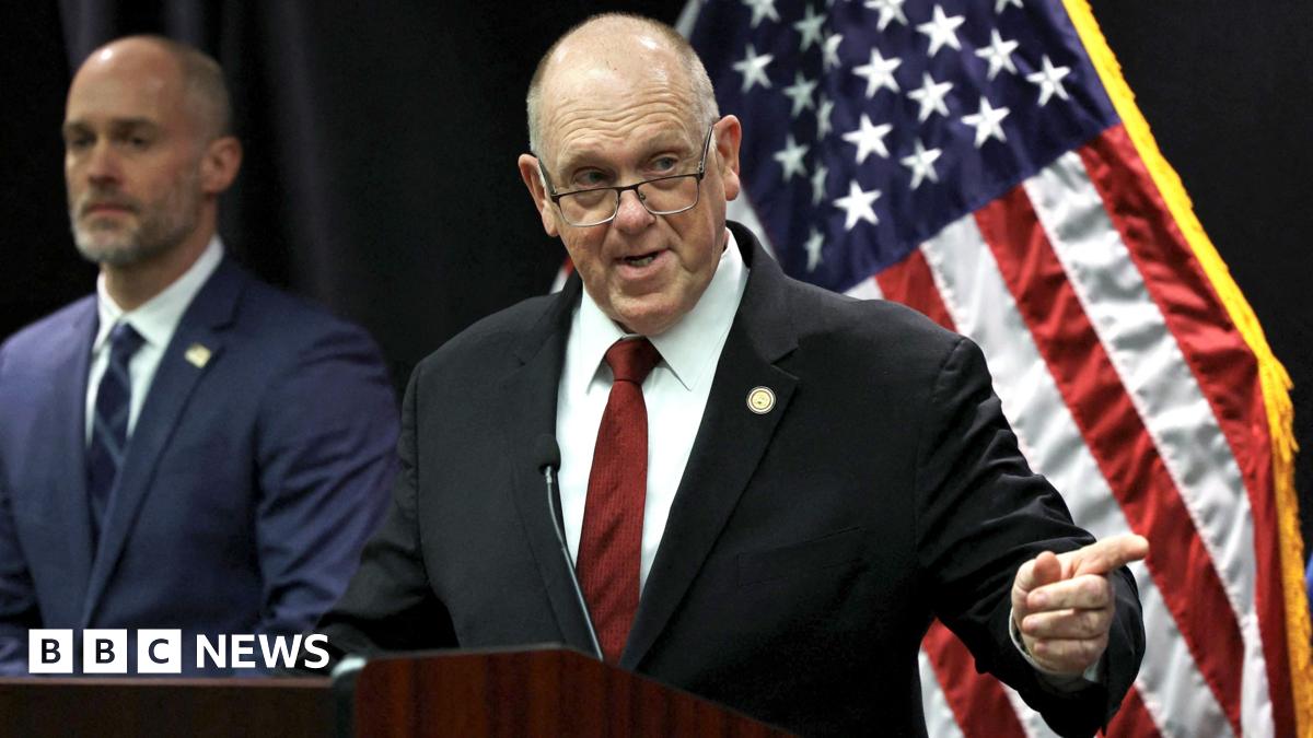 Tom Homan in front of American flag and a bald man in a blue suit stands at a lectern and points off to his left, while wearing a black suit, red tie and glasses that appear to be sliding down his nose