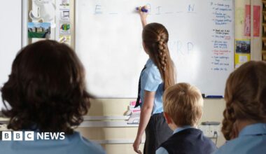 A girl in a light bue shirt and tie writes on a whiteboard. Other students are looking at the board.
