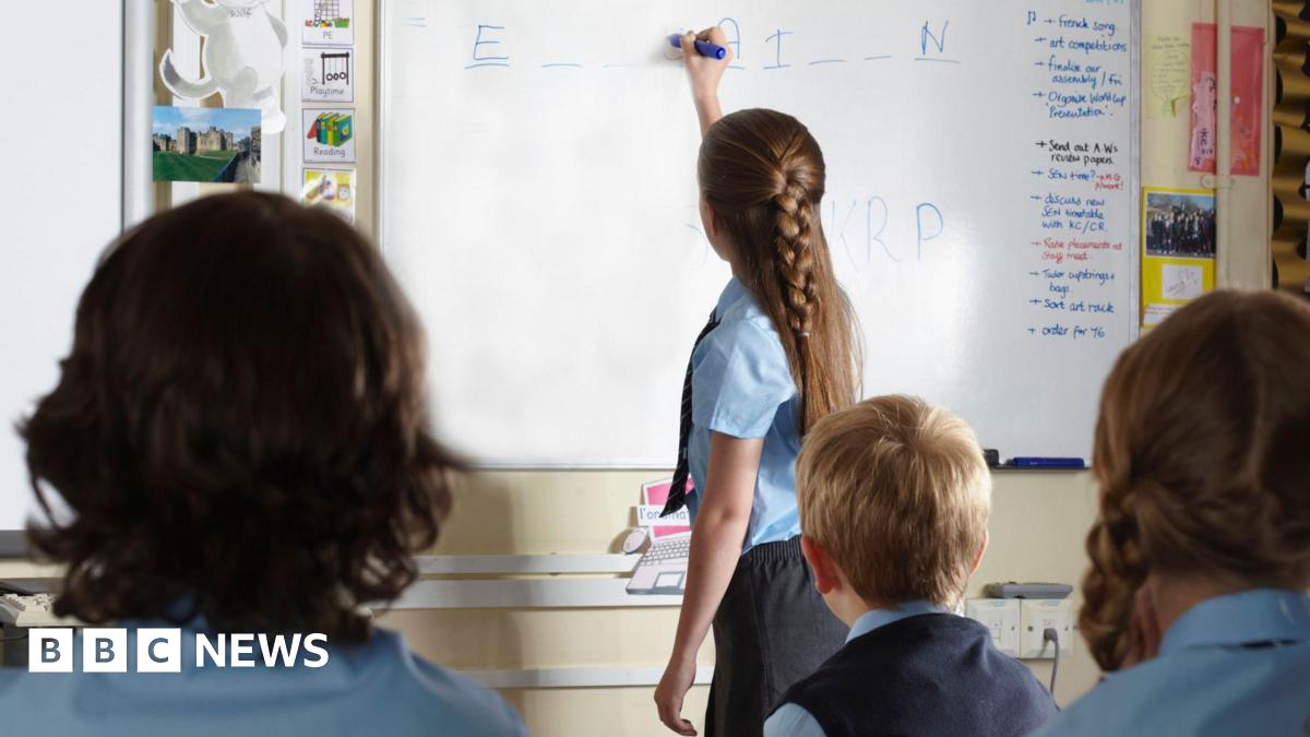 A girl in a light bue shirt and tie writes on a whiteboard. Other students are looking at the board.