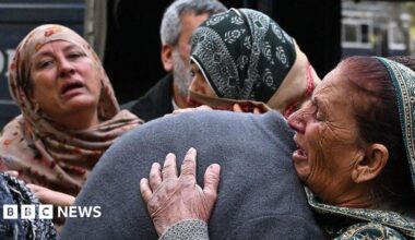 A group of people mourn the death of their relatives following a suicide bombing at a mosque in Islamabad. The woman are wearing head scarves and look distraught. One woman is hugging someone in a grey jumper, their head is obscured by her embrace.