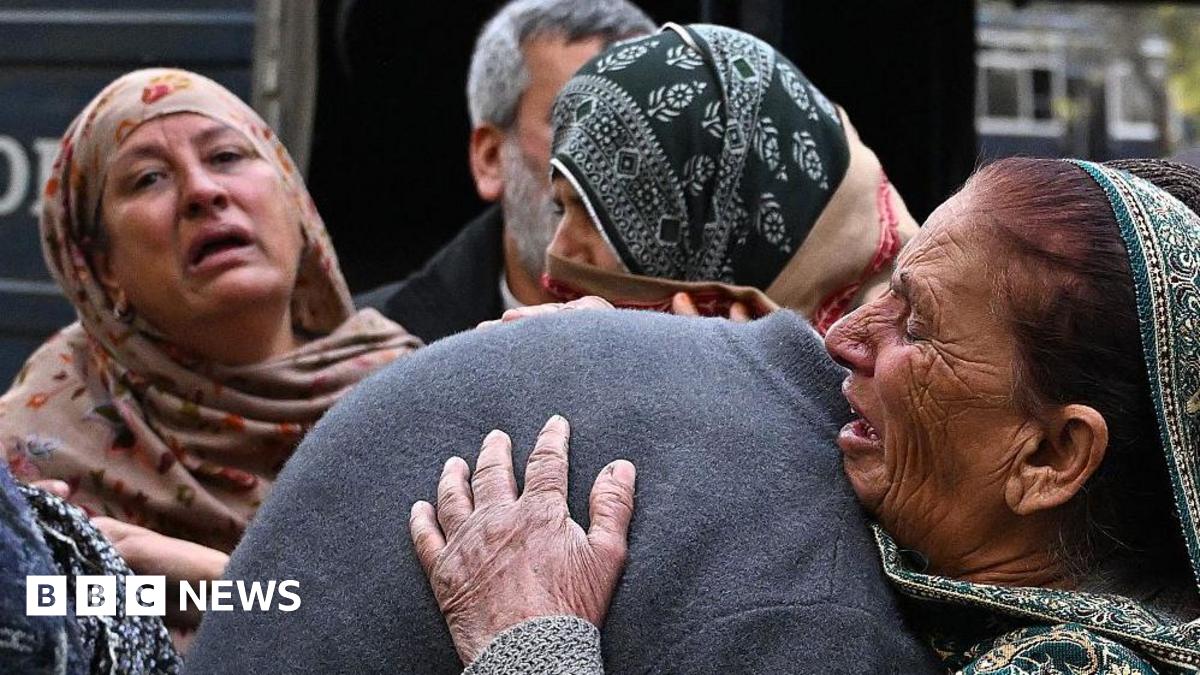 A group of people mourn the death of their relatives following a suicide bombing at a mosque in Islamabad. The woman are wearing head scarves and look distraught. One woman is hugging someone in a grey jumper, their head is obscured by her embrace.