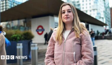Beth, in a light pink coat, stands at the entrance of the Battersea Power Station Underground station in London. Behind her are modern glass buildings and the tall white chimneys of the redeveloped Battersea Power Station.