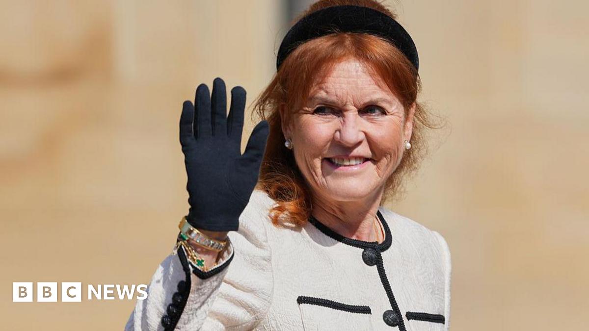 Sarah Ferguson waves her black-gloved hand while wearing a black headband, a cream jacket with a black trim, and large pearl earrings.