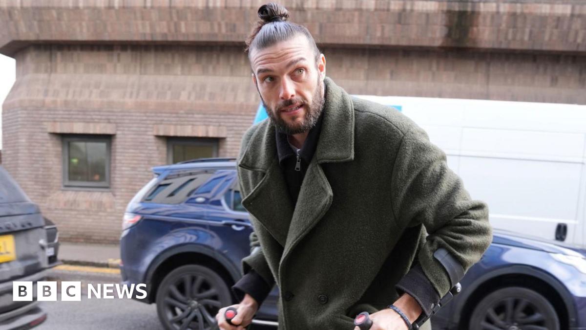 Andy Carroll has his brown hair tightly tied in a bun. He has a beard and is wearing a fluffy green jacket over a zipped shirt. He grips onto black crutches with both hands.