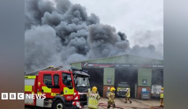 A thick, grey plume of smoke rises from a green, metal industrial warehouse. There is a fire engine parked out front and firefighters walking in front of the building.