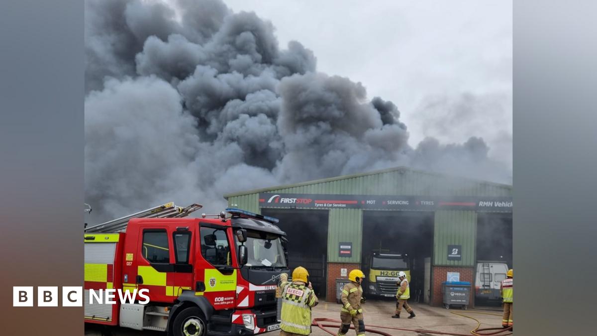 A thick, grey plume of smoke rises from a green, metal industrial warehouse. There is a fire engine parked out front and firefighters walking in front of the building.