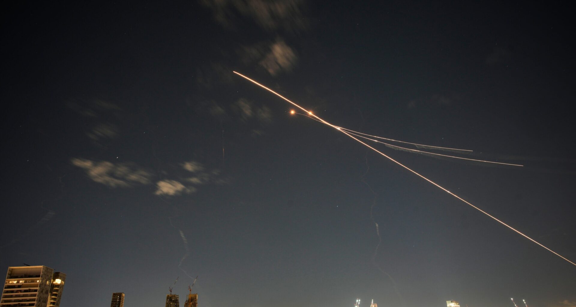Long-exposure photo of rockets and interceptors crossing the night sky above a city skyline with illuminated buildings.