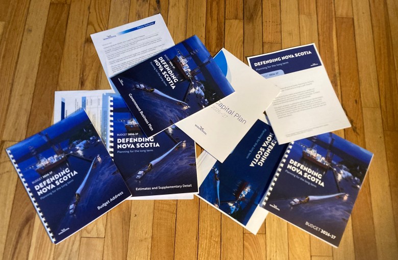 Five blue-coloured books sit on a hardwood floor. They are all titled "Defending Nova Scotia" but have different sub-headlines: Budget Address; Estimates and Supplmentary Detail; ;Budget 2026-27; Government Businss Plan; and Capital Plan.