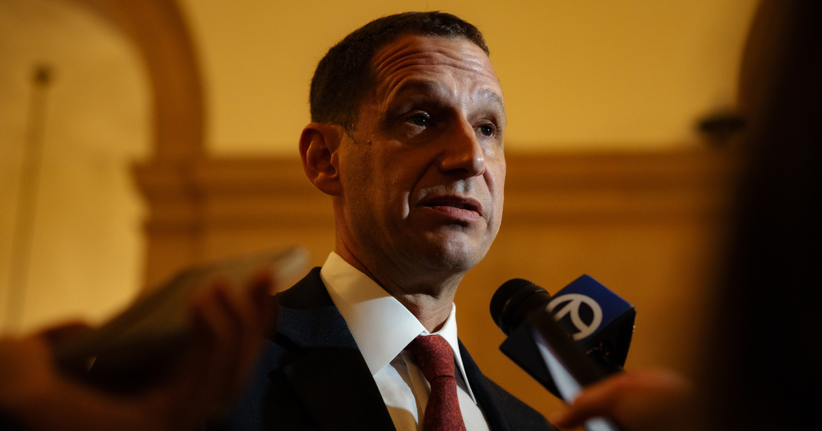 A man in a suit and red tie speaks into microphones, with a golden arch and warm lighting in the background.