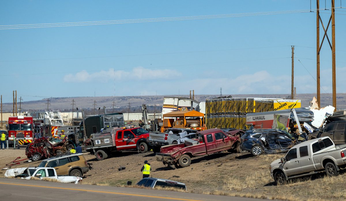 Multiple damaged vehicles, emergency responders, and trucks are shown at the scene of a major crash on the side of a rural road under clear skies.