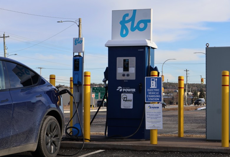 A blue car is plugged in and charging at an electric vehicle charging station. There are signs with instructions on how to user the charger.