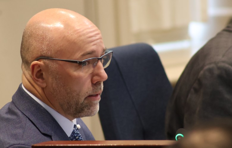 A white man with with a shaved head and closely trimmed beard and moustache in a grey-navy blazer, white shirt, and blue pattern tie.