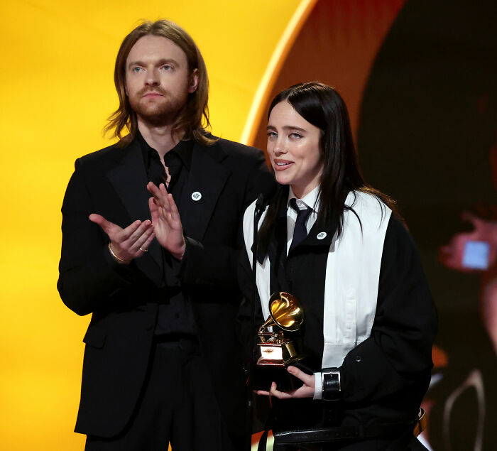 Billie Eilish holding a Grammy award on stage with a companion, amid critics discussing her lavish lifestyle.