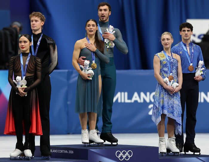 Olympian on podium holding medals and stuffed mascots as judge's scores spark cheating accusations controversy.