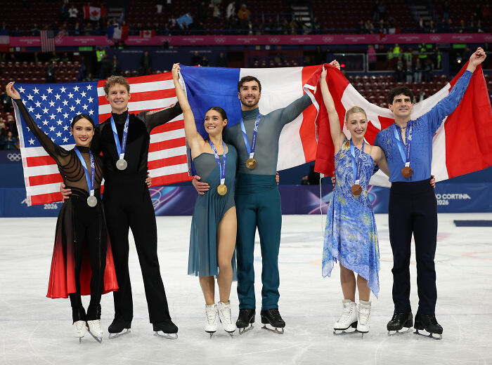 Olympian celebrating with medals on ice, holding national flags amid judge's conspicuous scores and cheating accusations.