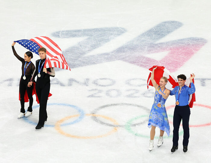 Two pairs of Olympic figure skaters celebrating on ice, holding national flags after medal awards in 2022 Winter Olympics.