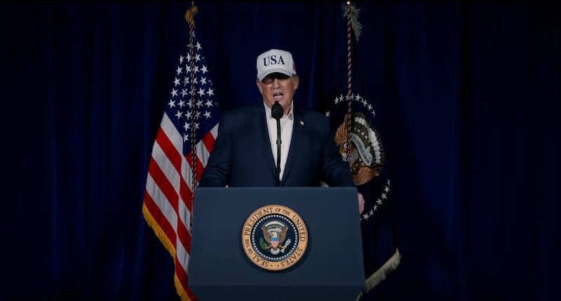 Donald Trump behind a lectern with the presidential seal. He is wearing a blue suit jacket, open-necked white shirt and white USA Trucker hat. He has behind him a U.S. and a military flag. He was speaking to launch his war on Iran.
