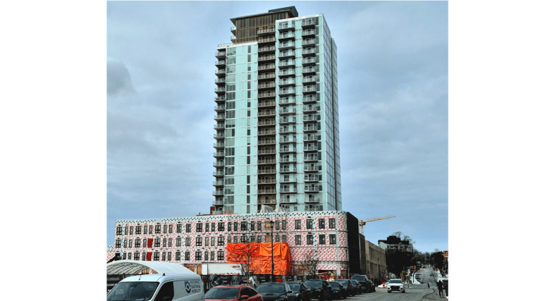 A 24-storey tower with greenish glass plating rises above a four-storey pedestal with red paper and an orang tarp. Cars line the street alongside the structure.