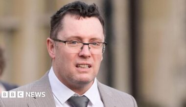 Image shows man in a light-brown suit with a silver tie and black-rimmed glasses. He has black hair and grey stuble.