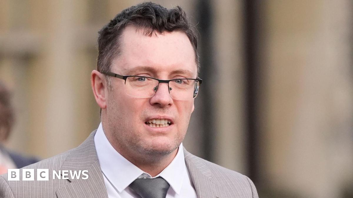 Image shows man in a light-brown suit with a silver tie and black-rimmed glasses. He has black hair and grey stuble.