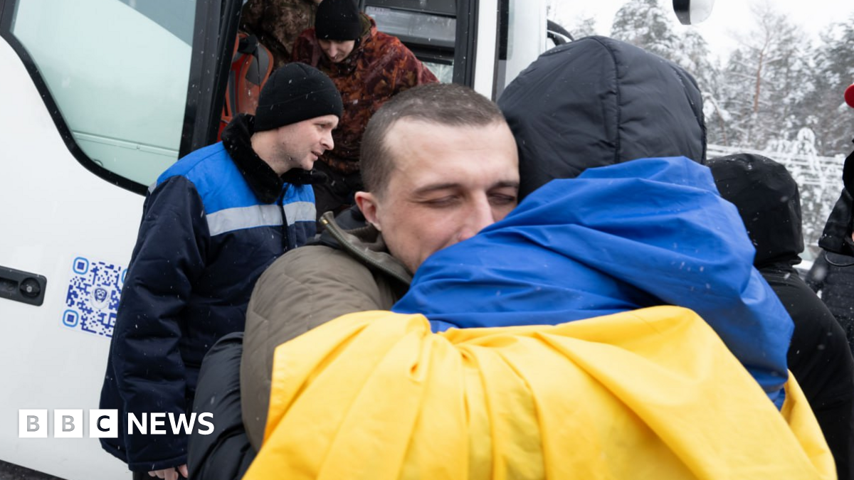 A Ukrainian soldier hugs a person draped in Ukraine's blue-and-yellow national flag after a prisoner swap with Russia. Other exchanged Ukrainian soldiers are seen leaving a bus in the background. Photo: 5 February 2026
