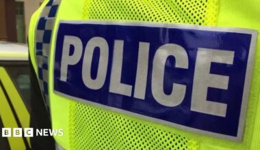 A tightly cropped view of a police vest. Police is spelt in white on a blue background. A police car is to the left.
