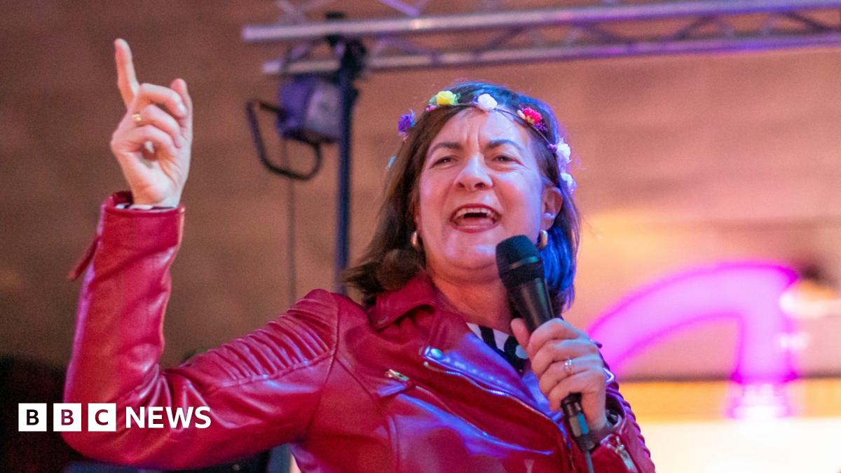 Eluned Morgan, a woman with short brown hair wearing a red leather jacket and has a crown of flowers on her head, sings into a microphone