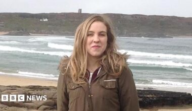 Natalie McNally. She has long blonde hair, wearing a long khaki coat and standing on a beach.