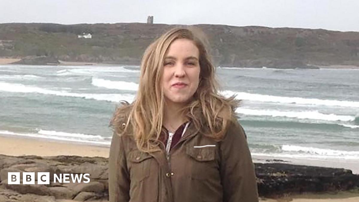 Natalie McNally. She has long blonde hair, wearing a long khaki coat and standing on a beach.