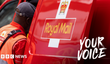 A Royal Mail postal worker dressed in a orange high-vis vest and red polo and navy blue cap looks into his Royal Mail work van side door