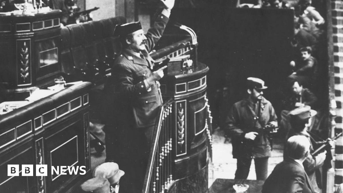 Coup leader Antonio Tejero Molina in police uniform stands at the top of stairs beside a lectern in the Spanish parliament brandishing a handgun in his right hand and with his left hand raised. Other armed men stand around him and in the distance MPs can be seen cowering down.
